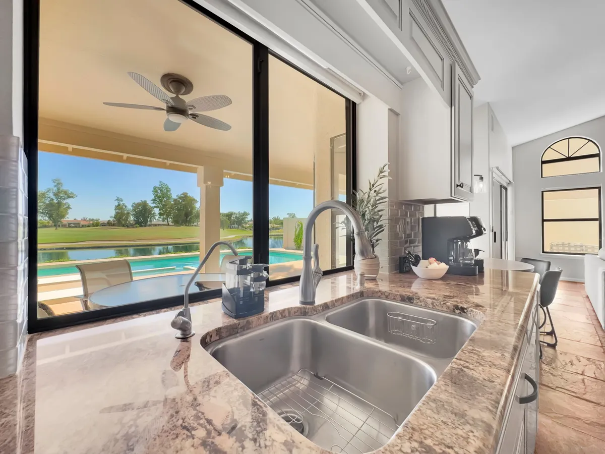 This interior kitchen view features a granite countertop, sink, and large windows that open onto an outdoor patio area, hinting at seamless indoor-outdoor living. Bright natural light fills the space, and the view connects the kitchen to the patio beyond.