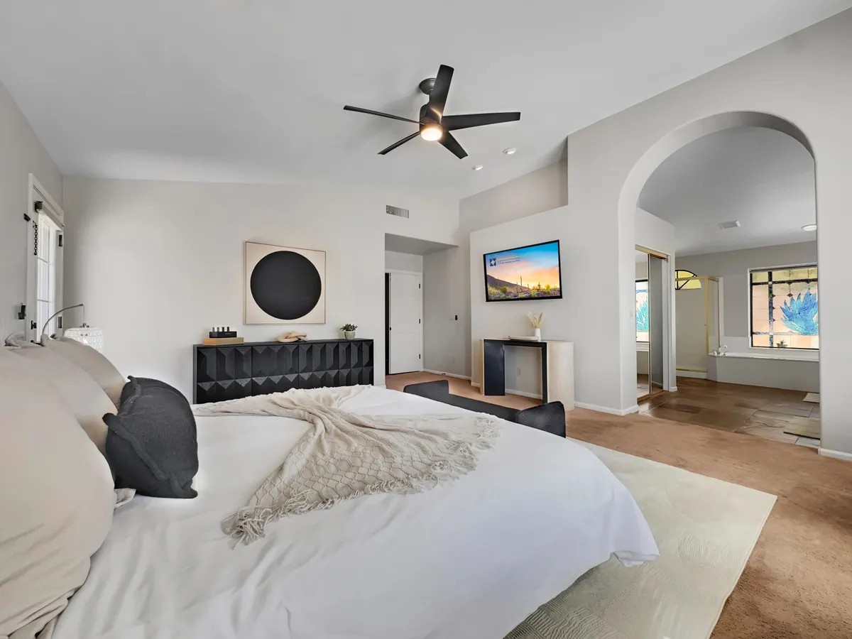 A serene bedroom with a king-sized bed dressed in gray and white bedding, a black tufted headboard, modern ceiling fan, TV mounted on the wall, and a large window with decorative glass. Soft natural light bathes the room, highlighting the calm atmosphere and inviting setting for a restful stay.