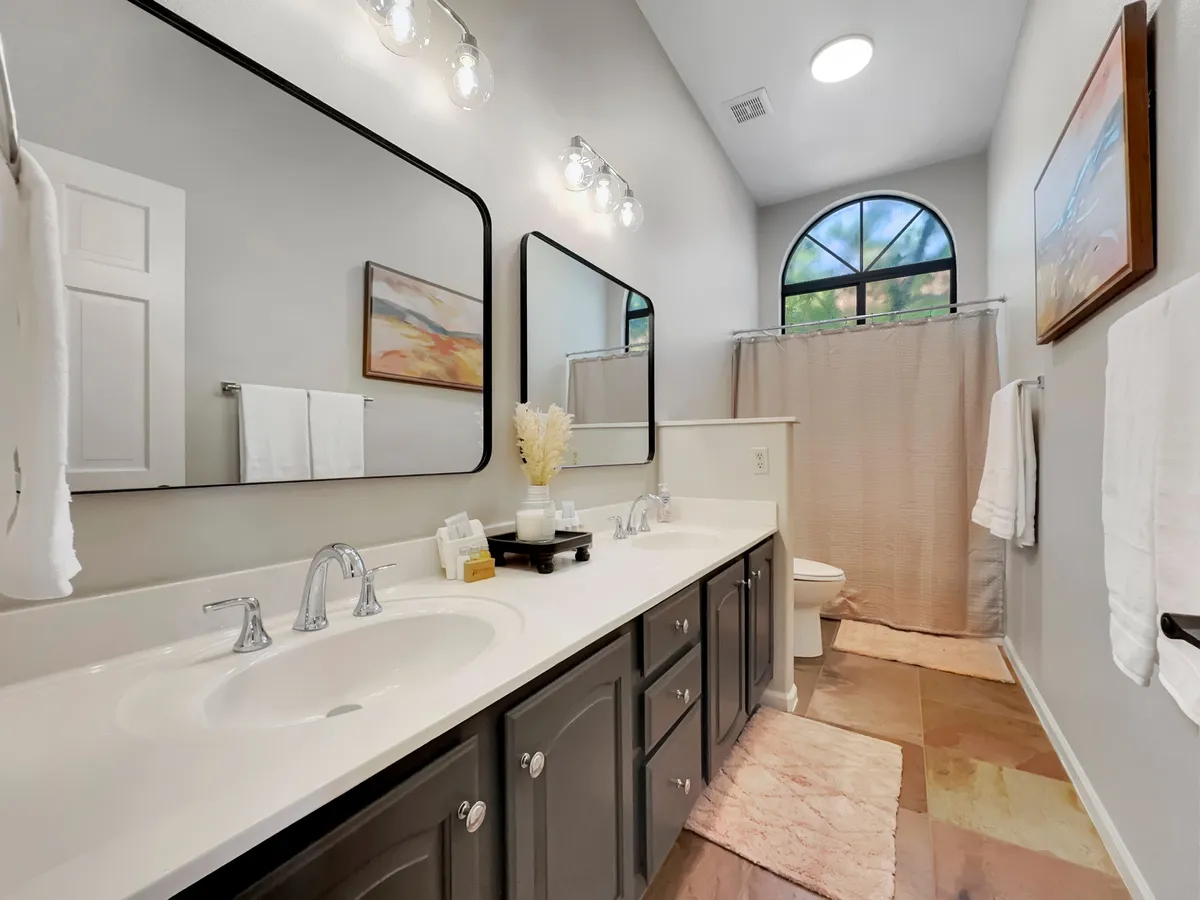 A refreshing bathroom with a double vanity, large mirrors, and an arched window letting in ample natural light. Decorative artwork adds a touch of elegance, making the space ideal for morning routines and quiet relaxation.