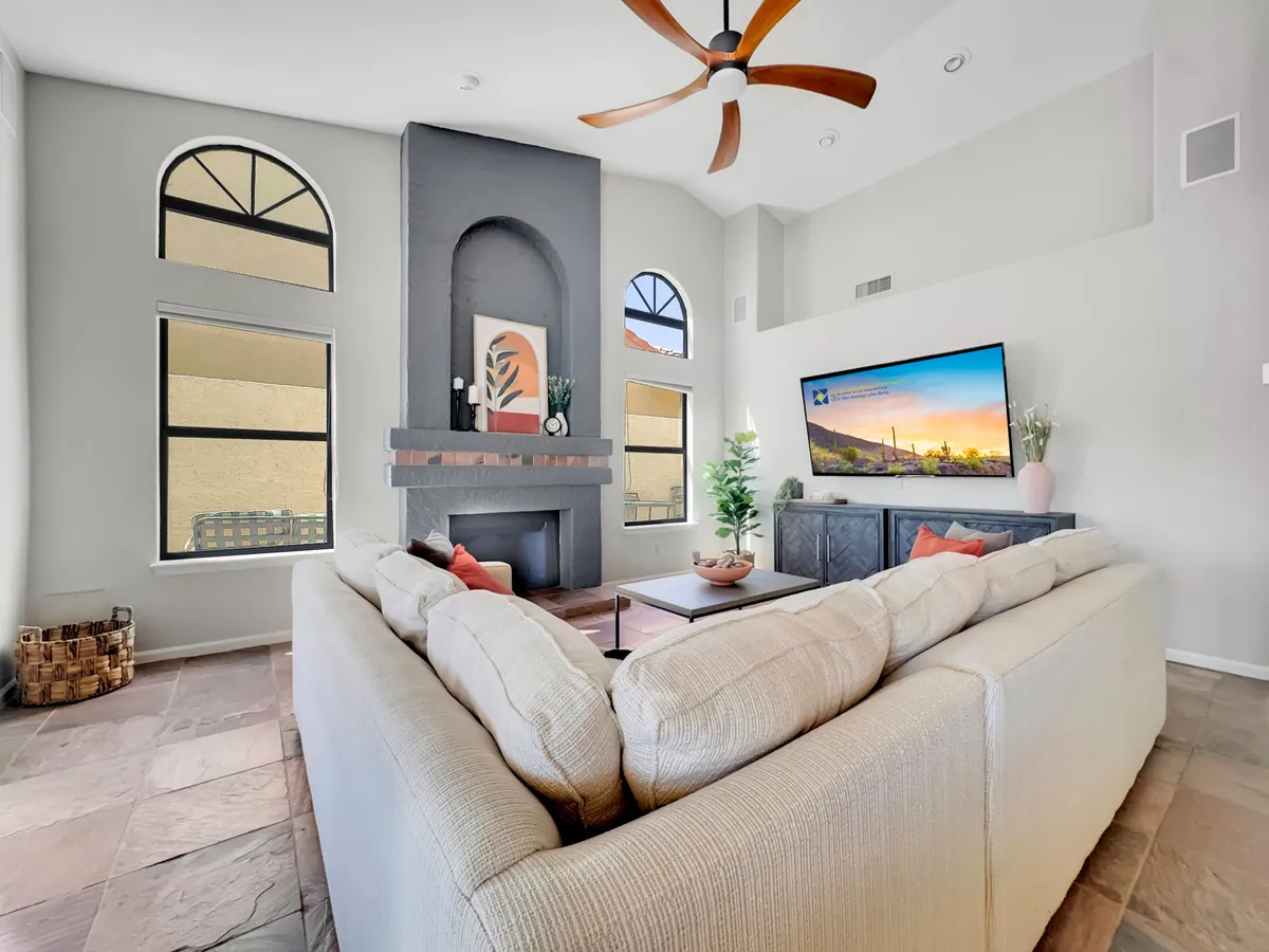 This image showcases a spacious living area with a large sectional sofa, a fireplace as a cozy focal point, a television with a scenic view displayed, and a decorative plant adding warmth. The setting is bright and filled with natural light streaming through large windows, giving a welcoming and communal atmosphere.