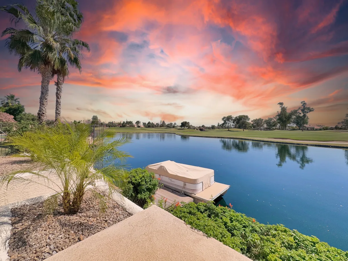 An inviting lakefront view framed by palm trees and landscaped gardens, with a boat covered by a tarp at the water's edge and a picturesque sunset sky. The shoreline suggests opportunities for boating, fishing, and peaceful family moments by the water.