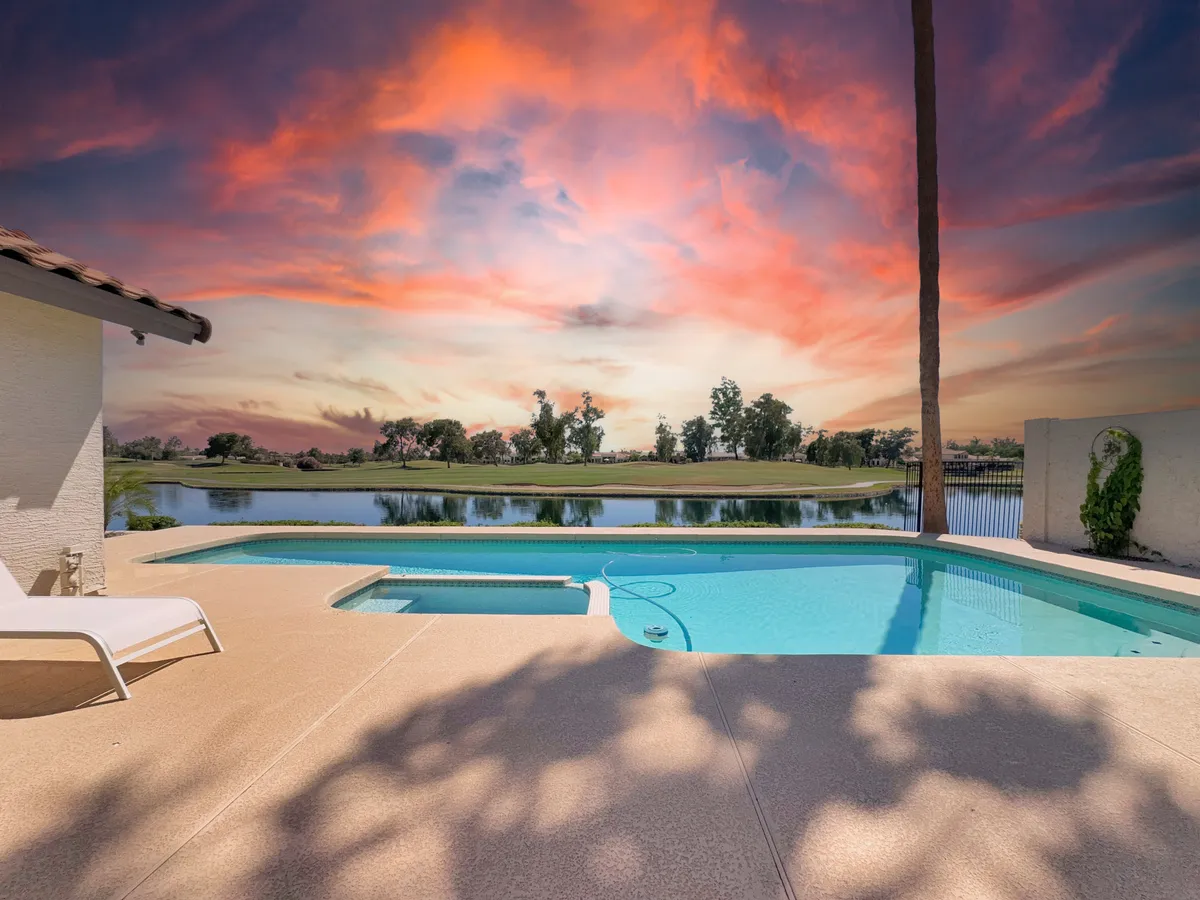 A stunning backyard scene featuring a sparkling swimming pool surrounded by lush greenery, with a distant lake and vibrant sunset sky completing the tranquil atmosphere. The space is clearly designed for family relaxation, with plenty of room for lounging and enjoying the water.