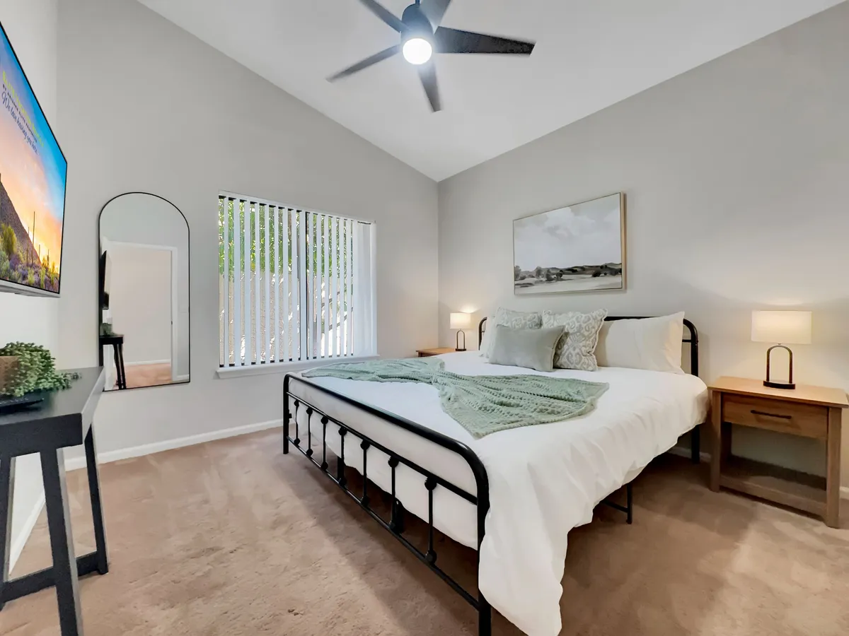 A calm bedroom scene with a cozy bed topped with a decorative throw, a mirror, TV, and nightstand with a lamp. Sunlight streams in, emphasizing the room's tranquil and inviting atmosphere, perfect for a restful night's sleep or quiet moments during the day.