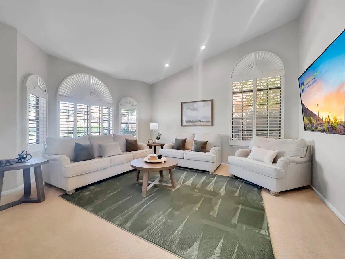A spacious, cozy living area featuring comfortable sofas arranged around a circular coffee table, large windows with shutters, and a television mounted on the wall. The space is ideal for socializing, relaxing, or enjoying games with family and friends.